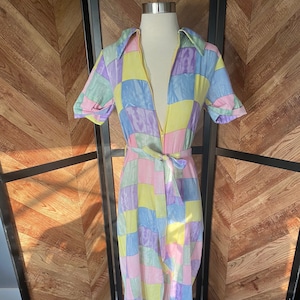 Può includere: Una tuta a maniche lunghe con un motivo a quadri in tonalità di rosa, giallo, verde e viola. La tuta ha una chiusura a zip e una cintura.