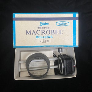 May include: A blue and white box with the text "Spirotone "Rapid-rail" MACROBEL Bellows for NIKON No. 500180" and a black and silver bellows camera attachment inside.