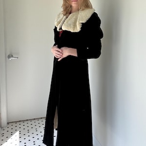 May include: A long black coat with a cream-coloured faux fur collar. The coat is floor-length and has a slit up the front. The woman is standing in front of a white door and wall.