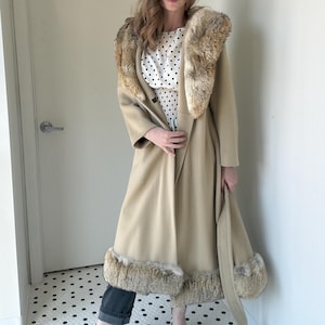 Peut inclure: Un long manteau beige avec un col en fourrure et une garniture en fourrure aux poignets et à l'ourlet. Le manteau est ouvert, révélant une robe à pois. Le manteau a une ceinture assortie. Le modèle se tient devant une porte blanche.