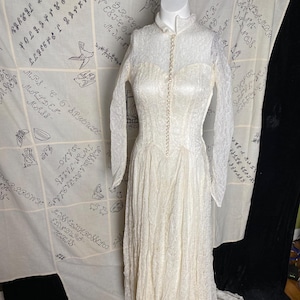 Könnte beinhalten: Ein weißes Spitzen-Hochzeitskleid mit langem Schleier und hohem Ausschnitt. Das Kleid hat eine Reihe von Perlmuttknöpfen auf der Vorderseite.