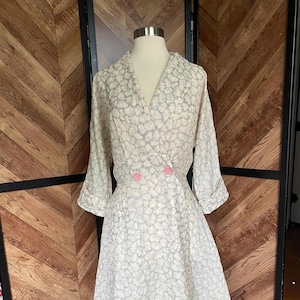 May include: A white dress with a floral pattern and pink buttons. The dress has a V-neckline and long sleeves.