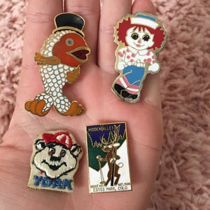 May include: Four enamel pins featuring a red fish wearing a top hat, a red-haired doll wearing a blue dress, a cartoon squirrel wearing a red hat, and a cartoon deer wearing a red hat and holding skis. The deer pin has the text "HIDDEN VALLEY" and "ESTES PARK, COLO."