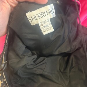 Pu&ograve; includere: Un abito nero Sherri Hill con un'etichetta che recita "SHERRI HILL" e "6 TER". L'abito ha una fodera nera e dettagli in pizzo. L'etichetta indica anche "MADE IN CHINA".