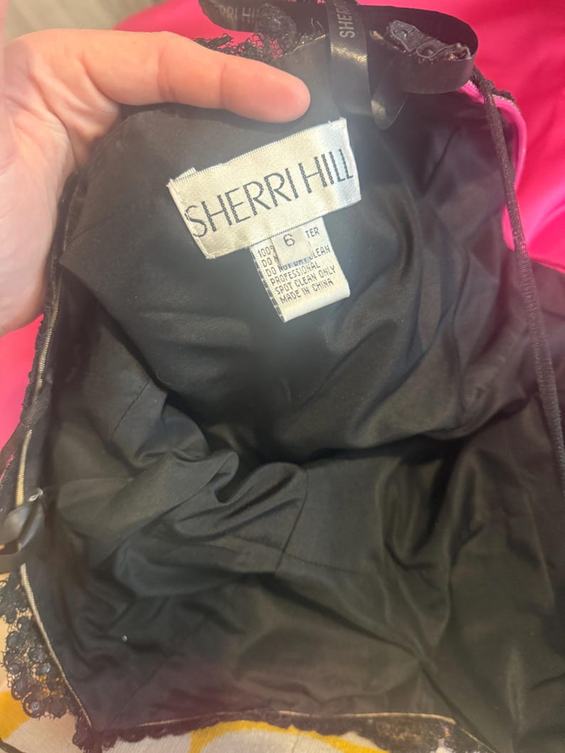 Pu&ograve; includere: Un abito nero Sherri Hill con un'etichetta che recita "SHERRI HILL" e "6 TER". L'abito ha una fodera nera e dettagli in pizzo. L'etichetta indica anche "MADE IN CHINA".