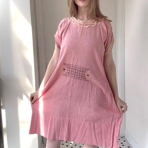 May include: A light pink dress with a round neckline and short sleeves. The dress features a decorative embroidered design on the front and a ruffled hem. The neckline and sleeve edges are trimmed with a cream-colored lace.