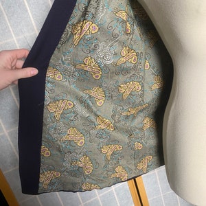 Puede incluir: El interior de una chaqueta azul marino con un forro estampado en verde y amarillo. El patr&oacute;n presenta mariposas y remolinos.