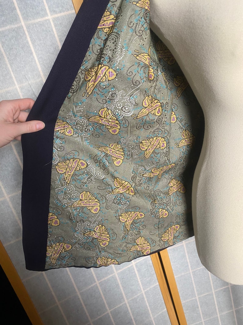 Puede incluir: El interior de una chaqueta azul marino con un forro estampado en verde y amarillo. El patr&oacute;n presenta mariposas y remolinos.