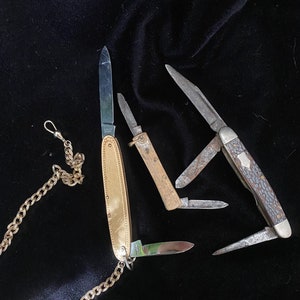 May include: Three pocket knives with different designs and finishes. One knife has a gold-coloured handle and a chain attached to it. The other two knives have a more traditional design with a brown handle and a silver blade.