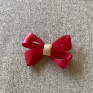 Könnte beinhalten: Eine rosafarbene Schleife aus Plastik mit einer weißen Mitte. Die Schleife liegt auf einer strukturierten grauen Oberfläche.
