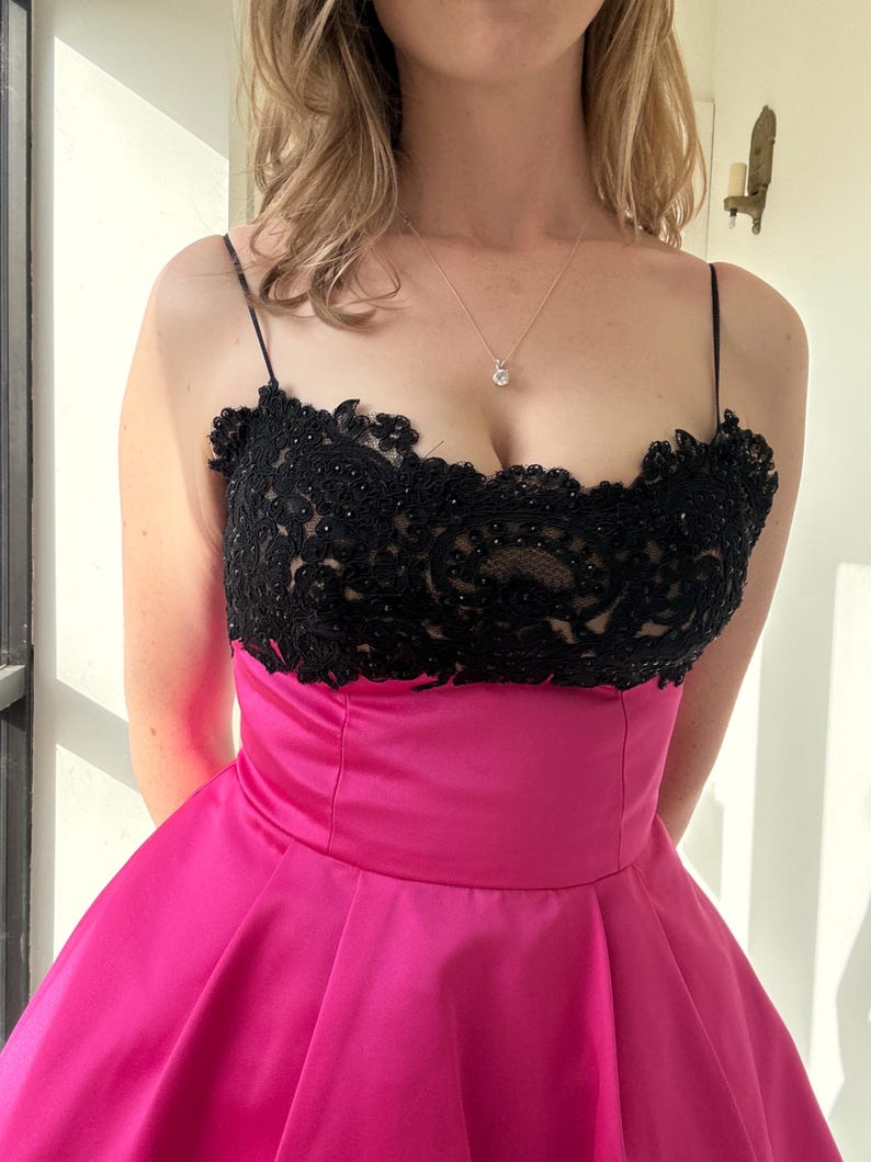 Pu&ograve; includere: Un abito da sera con corpetto in pizzo nero e gonna rosa acceso. Il corpetto ha sottili spalline e intricate perline. La gonna &egrave; di un rosa brillante e tinta unita, creando un contrasto sorprendente. &Egrave; visibile anche una collana d'argento con un piccolo pendente.