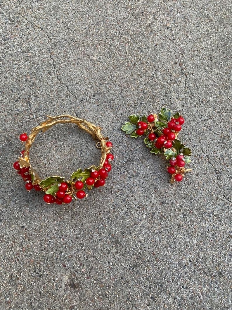 May include: A gold bracelet and brooch set with red berries and green leaves. The jewelry is made of metal and has a vintage style.