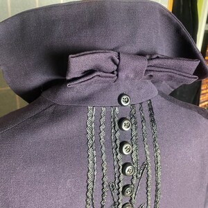 Puede incluir: Una chaqueta de color morado oscuro con un lazo negro en el cuello y un ribete bordado negro en la espalda. El ribete presenta una serie de botones negros.