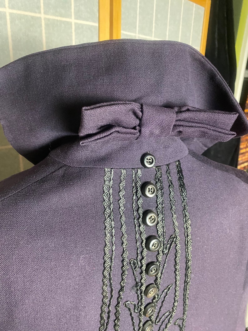 Puede incluir: Una chaqueta de color morado oscuro con un lazo negro en el cuello y un ribete bordado negro en la espalda. El ribete presenta una serie de botones negros.