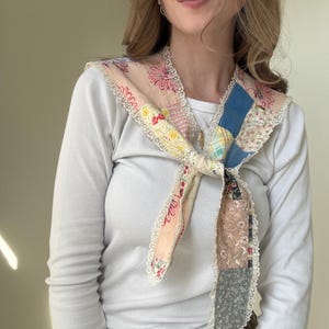Pode incluir: Um cachecol de patchwork com uma variedade de tecidos florais e estampados em rosa, azul, amarelo e cinza. O cachecol é adornado com renda branca e amarrado no pescoço. Usado sobre uma camisa branca de manga comprida.