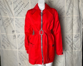 Puede incluir: Una chaqueta roja con cremallera, cinturón negro y dos bolsillos. La chaqueta tiene cuello y está hecha de una tela suave y texturizada.