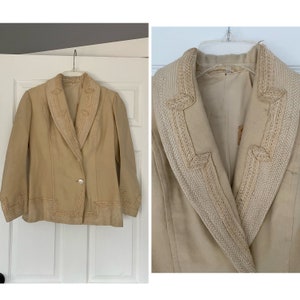 Puede incluir: Una chaqueta beige con un ribete blanco bordado alrededor del cuello y las mangas. La chaqueta tiene un cierre de botón único y un cuello puntiagudo.