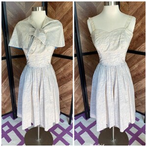 Pode incluir: Um vestido branco com um padrão de redemoinho azul e branco. O vestido tem um decote de coração e uma saia cheia. É combinado com uma jaqueta bolero branca com uma borda azul.