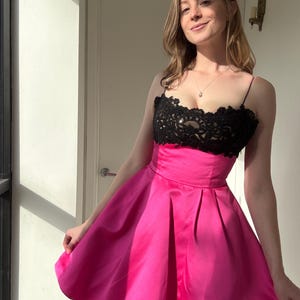 Pu&ograve; includere: Un abito rosa acceso con corpetto in pizzo nero. L'abito ha una gonna a pieghe e sottili spalline. L'illuminazione mette in risalto la trama del tessuto, creando un look vivace e accattivante.