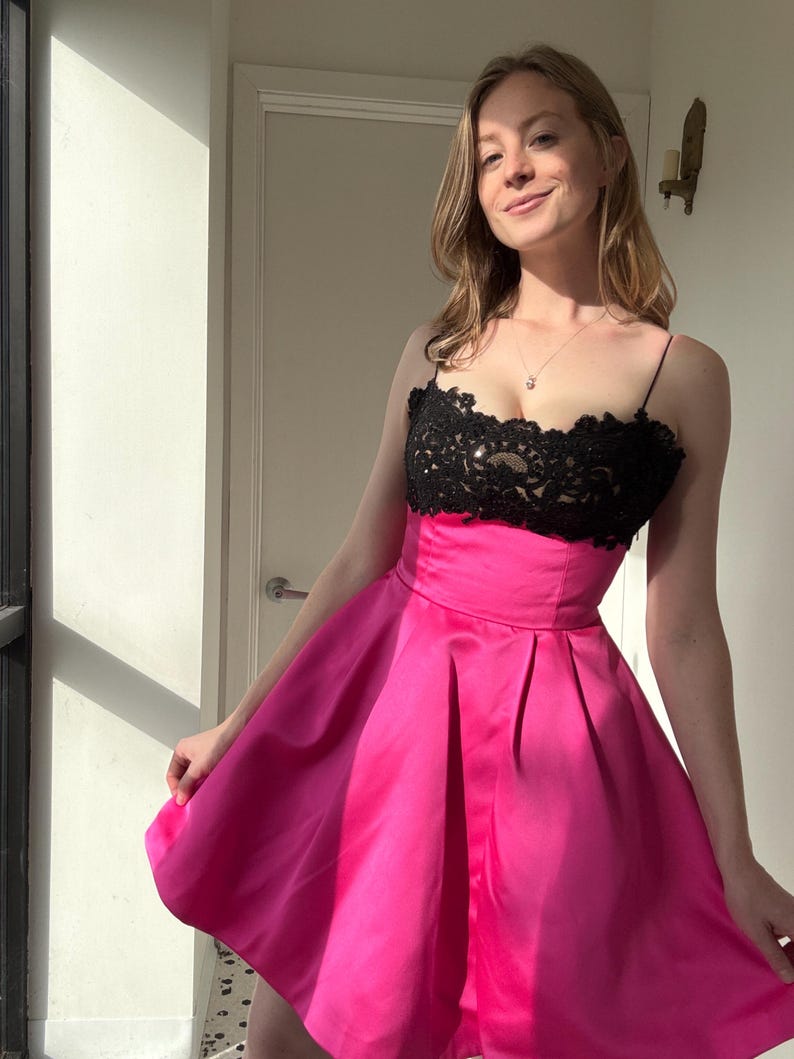 Pu&ograve; includere: Un abito rosa acceso con corpetto in pizzo nero. L'abito ha una gonna a pieghe e sottili spalline. L'illuminazione mette in risalto la trama del tessuto, creando un look vivace e accattivante.