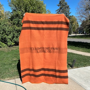 Puede incluir: Una manta de lana a rayas naranja y marrón con el texto "Yellowstone Park Lodge Co." impreso en negro.