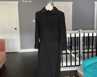 May include: A black skirt suit with a black fur collar. The suit is on a mannequin and is displayed on a striped rug.