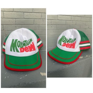 Puede incluir: Una gorra de camionero blanca, verde y roja con el logotipo de Mountain Dew. La gorra tiene una parte trasera de malla y una visera verde con un borde rojo.