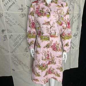 May include: A pink quilted robe with a floral print featuring a cottage scene. The robe has long sleeves and a button-up front.