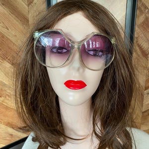May include: A pair of large, clear, plastic framed sunglasses with purple gradient lenses. The sunglasses are on a mannequin head with long brown hair and red lipstick.