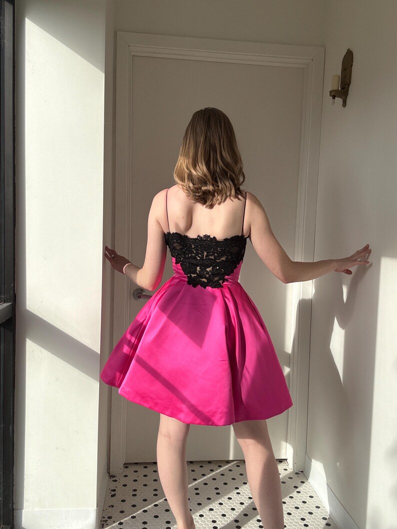 Pu&ograve; includere: Un abito rosa acceso con corpetto in pizzo nero e spalline sottili. La gonna &egrave; svasata e l'abito sembra essere realizzato in un materiale simile al raso. L'abito &egrave; lungo fino al ginocchio.