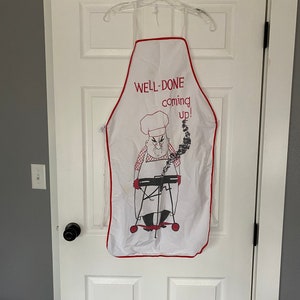 May include: White kitchen apron with a red trim. It features a cartoon chef with a grill and the text "WELL-DONE coming up!"