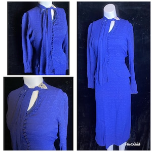 May include: A vintage cobalt blue two-piece dress set. The set includes a long-sleeved jacket with a keyhole neckline and a row of buttons, and a matching knee-length skirt. The fabric has a subtle textured pattern. The jacket has a decorative collar.