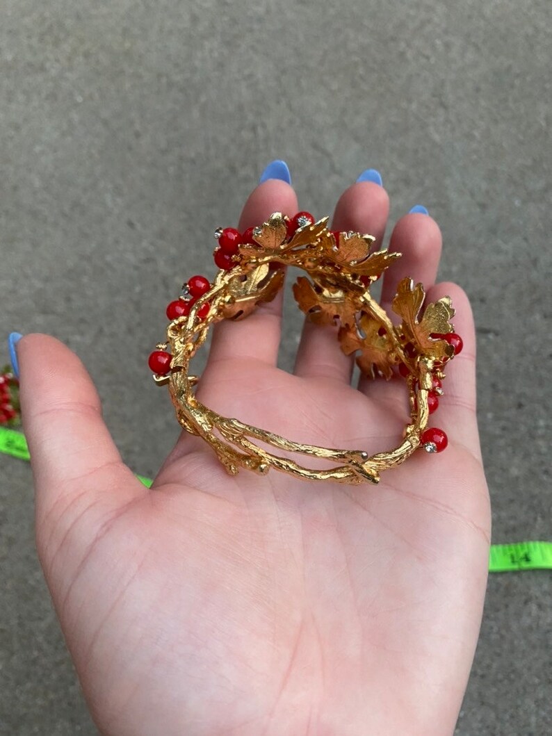 May include: Gold tone bracelet with red glass beads and gold leaf accents. The bracelet is open and has a delicate, twig-like design.