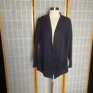 Puede incluir: Una chaqueta de punto azul oscuro de manga larga con un dise&ntilde;o bordado en negro. La chaqueta est&aacute; abierta por delante y tiene capucha.
