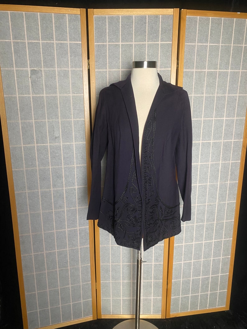 Puede incluir: Una chaqueta de punto azul oscuro de manga larga con un dise&ntilde;o bordado en negro. La chaqueta est&aacute; abierta por delante y tiene capucha.
