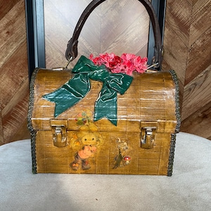 May include: A vintage wooden handbag with a brown leather handle and a green bow. The bag features a decorative floral arrangement and two metal clasps. The front of the bag has a cartoon illustration.