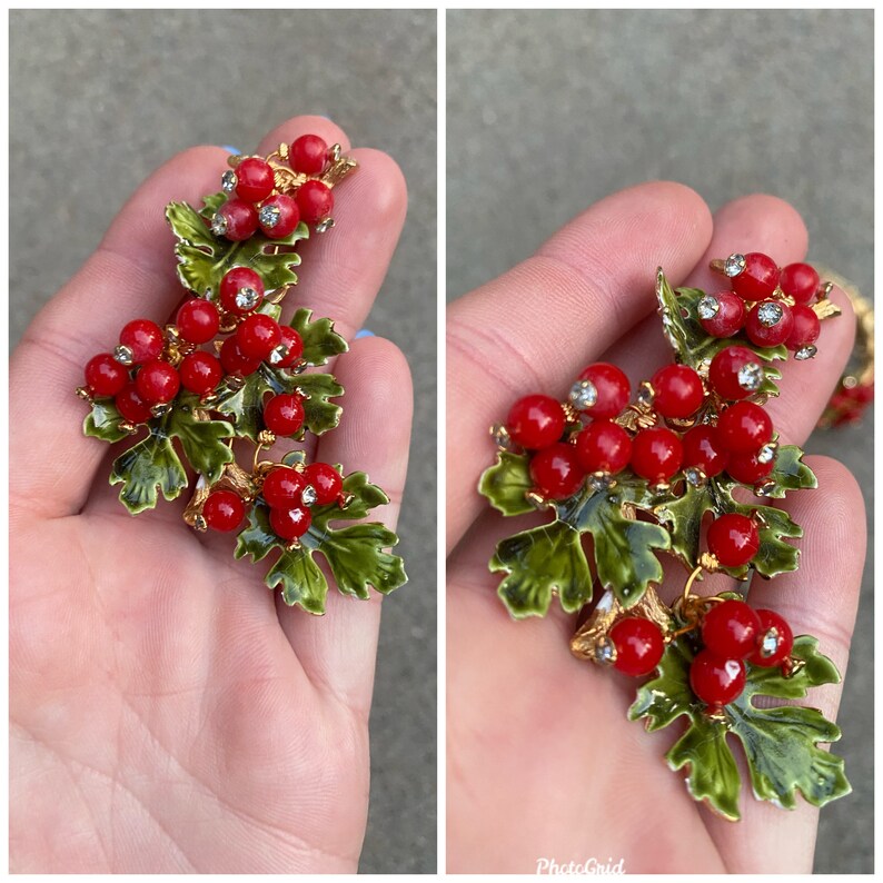 May include: A pair of red and green enamel berry brooch pins with rhinestones. The berries are clustered together on a branch with green leaves.