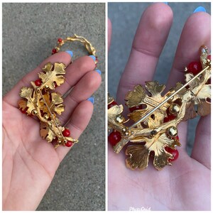 May include: Gold-toned brooch with red glass beads and a leaf design. The brooch has a pin back and is marked "Boucher" on the back.