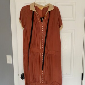 May include: A rust-coloured dress with a white collar and black ribbon ties. The dress has a pleated skirt and a row of buttons down the front.