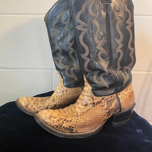 May include: A pair of cowboy boots with a snakeskin pattern on the foot and black leather uppers. The boots have decorative stitching and a pointed toe. The heels are black and angled.