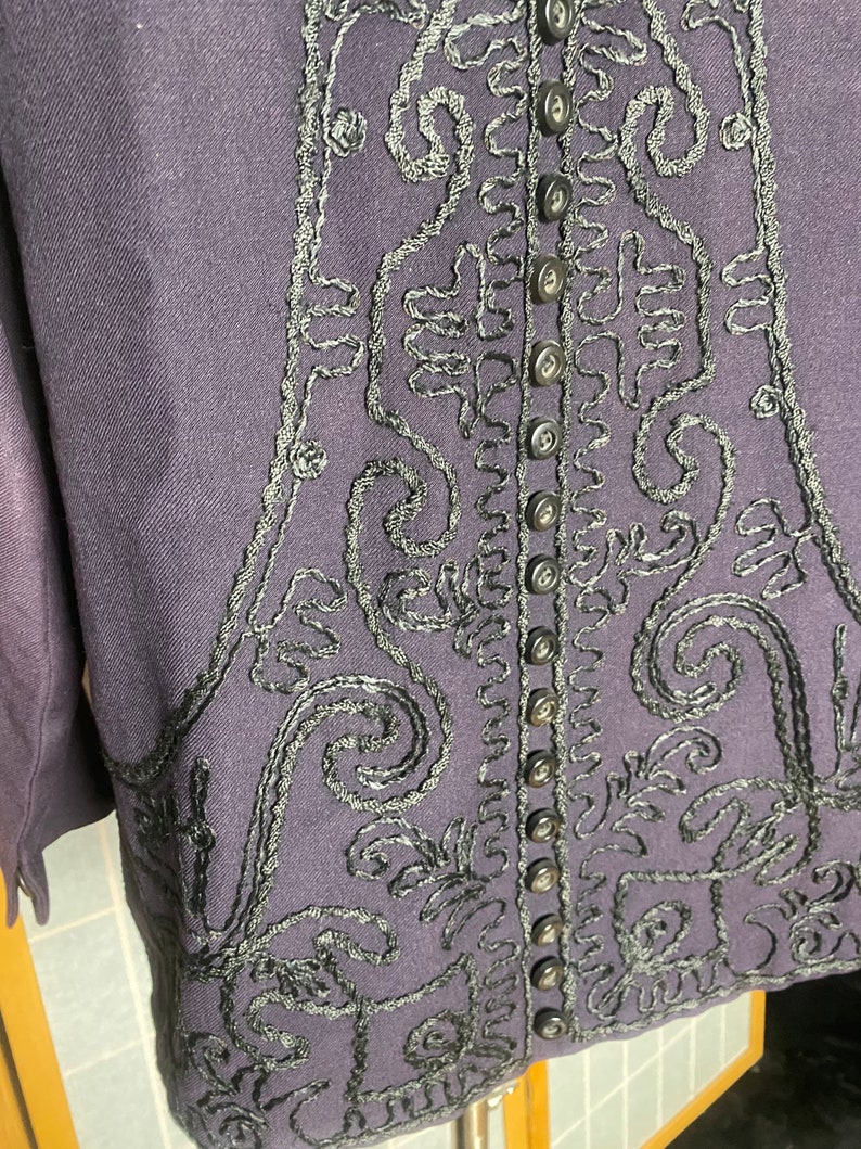 Puede incluir: Una chaqueta de color morado oscuro con detalles bordados en negro y una fila de botones negros en la espalda.
