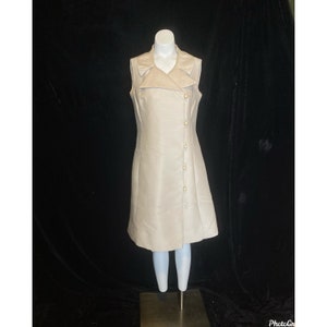 Puede incluir: Un vestido sin mangas de color beige claro con cuello y una fila de botones en la parte delantera. El vestido tiene una falda ligeramente acampanada y está hecho de una tela suave y sedosa.