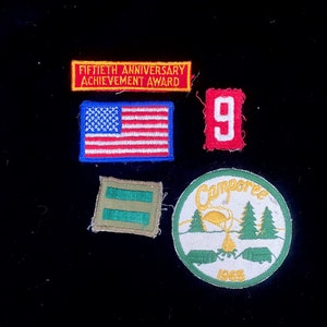 May include: Five embroidered patches, including a red, white, and blue American flag, a red patch with the number "9", a green patch with two horizontal stripes, a red patch with the text "Fiftieth Anniversary Achievement Award", and a white, green, and yellow patch with the text "Campore 1965" and a campfire scene.