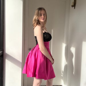 Pu&ograve; includere: Un vivace abito corto rosa acceso con corpetto in pizzo nero. L'abito presenta sottili spalline e una gonna ampia e svasata. La modella &egrave; in piedi davanti a una porta bianca e una finestra, con ombre.