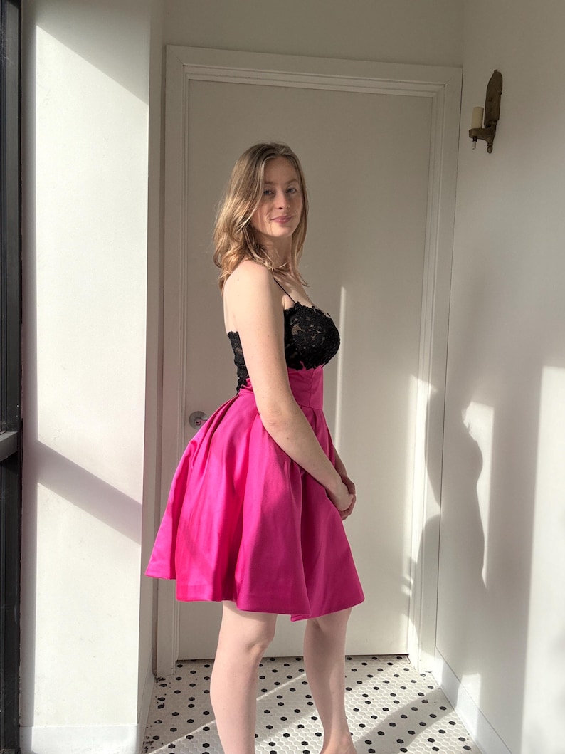 Pu&ograve; includere: Un vivace abito corto rosa acceso con corpetto in pizzo nero. L'abito presenta sottili spalline e una gonna ampia e svasata. La modella &egrave; in piedi davanti a una porta bianca e una finestra, con ombre.