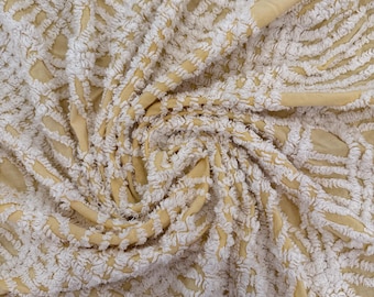 May include: Close-up of a textured fabric with a cream-colored, raised, geometric pattern over a pale yellow background. The fabric is soft and appears to be draped, with visible folds and shadows. The design creates a sense of depth and visual interest.