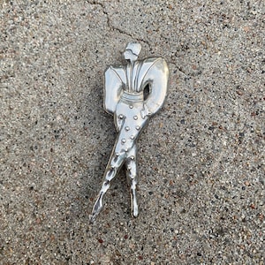 Könnte beinhalten: Silberne Brosche mit einer stilisierten Figur mit einem Tupfenmuster. Die Figur trägt ein langärmeliges Outfit mit übertriebenen Schultern.