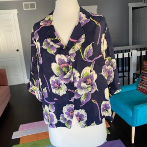 Könnte beinhalten: Eine marineblaue, halbtransparente Bluse mit einem Blumendruck mit weißen, lila und grünen Blumen. Die Bluse hat einen Kragen und einen Knopfverschluss vorne.