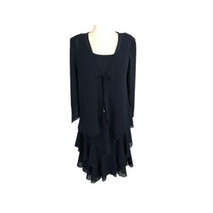 May include: A navy blue formal dress set. The set includes a sleeveless dress with a square neckline and a tiered skirt, and a matching long-sleeve jacket with a tie closure. The jacket has a scalloped hemline. The dress is displayed on a mannequin.