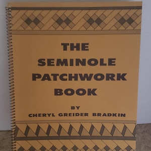 Peut inclure: Un livre à spirales intitulé "The Seminole Patchwork Book" par Cheryl Greider Bradkin. La couverture est beige avec des motifs géométriques et du texte bruns. Le livre est présenté sur un support noir.